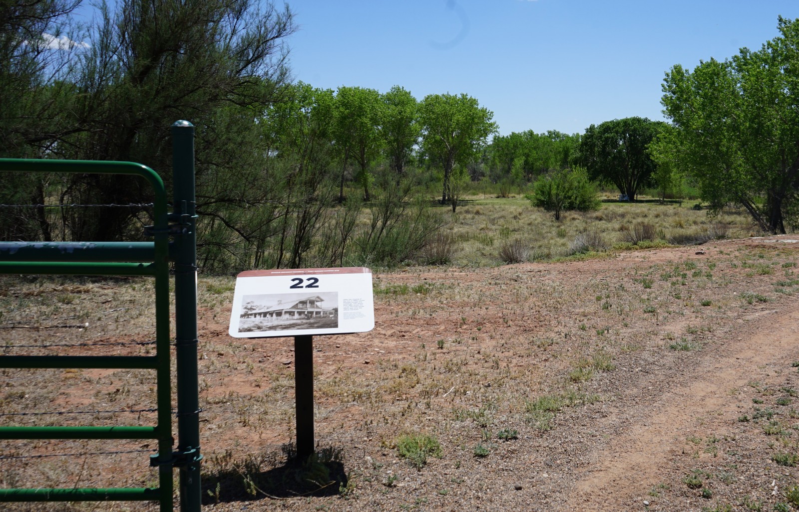 Bosque Redondo Memorial Audio Tour Stop 22 beside green gate, flood plain and trees in the foreground.