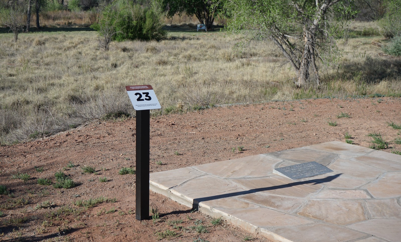 Bosque Redondo Memorial Audio Tour Stop 23 beside flagstone and Billy the Kid gravestone plaque, flood plain and trees in the foreground.