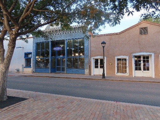 The exterior of Taylor-Mesilla Historic Site. A large tree frames the image on the left. The front fa?ade of the historic site is between the trees and across the street. The blue wood and large plate glass window fa?ade is on the left and two white painted double doors are to the left.
